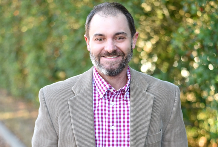 Professional man in gray blazer and pink checkered shirt smiling outdoors against green foliage background.
