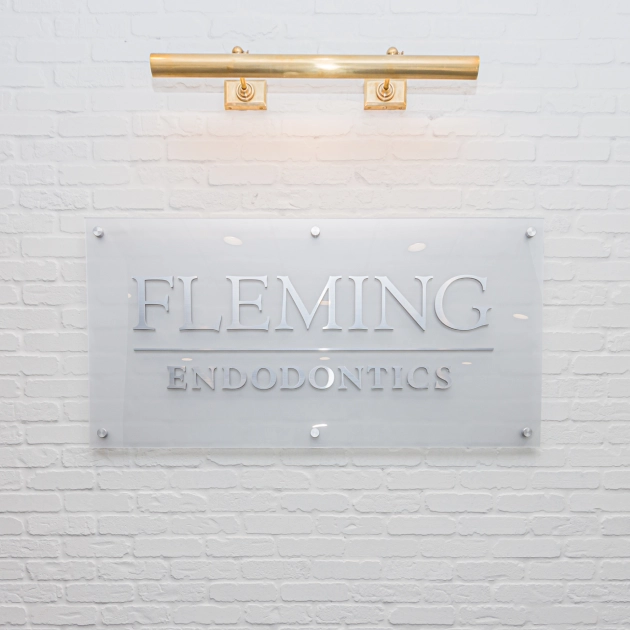 Frosted glass office sign reading "FLEMING ENDODONTICS" mounted on white brick wall with brass lighting fixture.