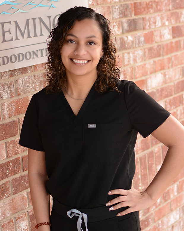 Woman in black scrubs smiling at camera against brick wall background at dental office.