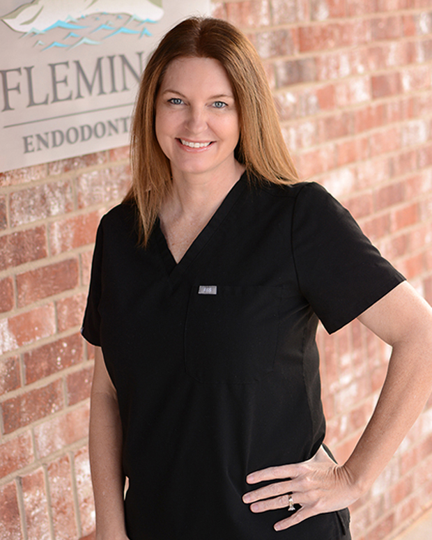 Woman in black scrubs smiling at camera in front of brick wall with Fleming Endodontic signage.