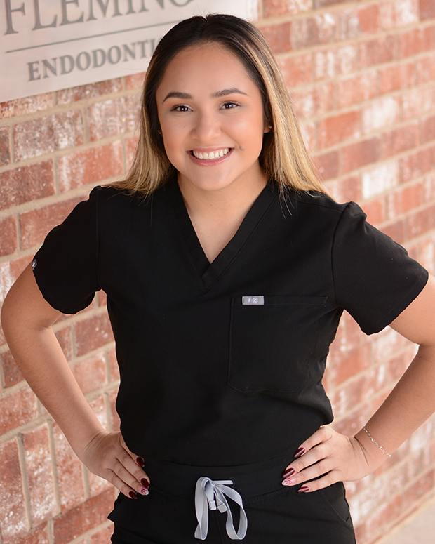 Female dental professional in black scrubs smiling at camera against brick wall background.